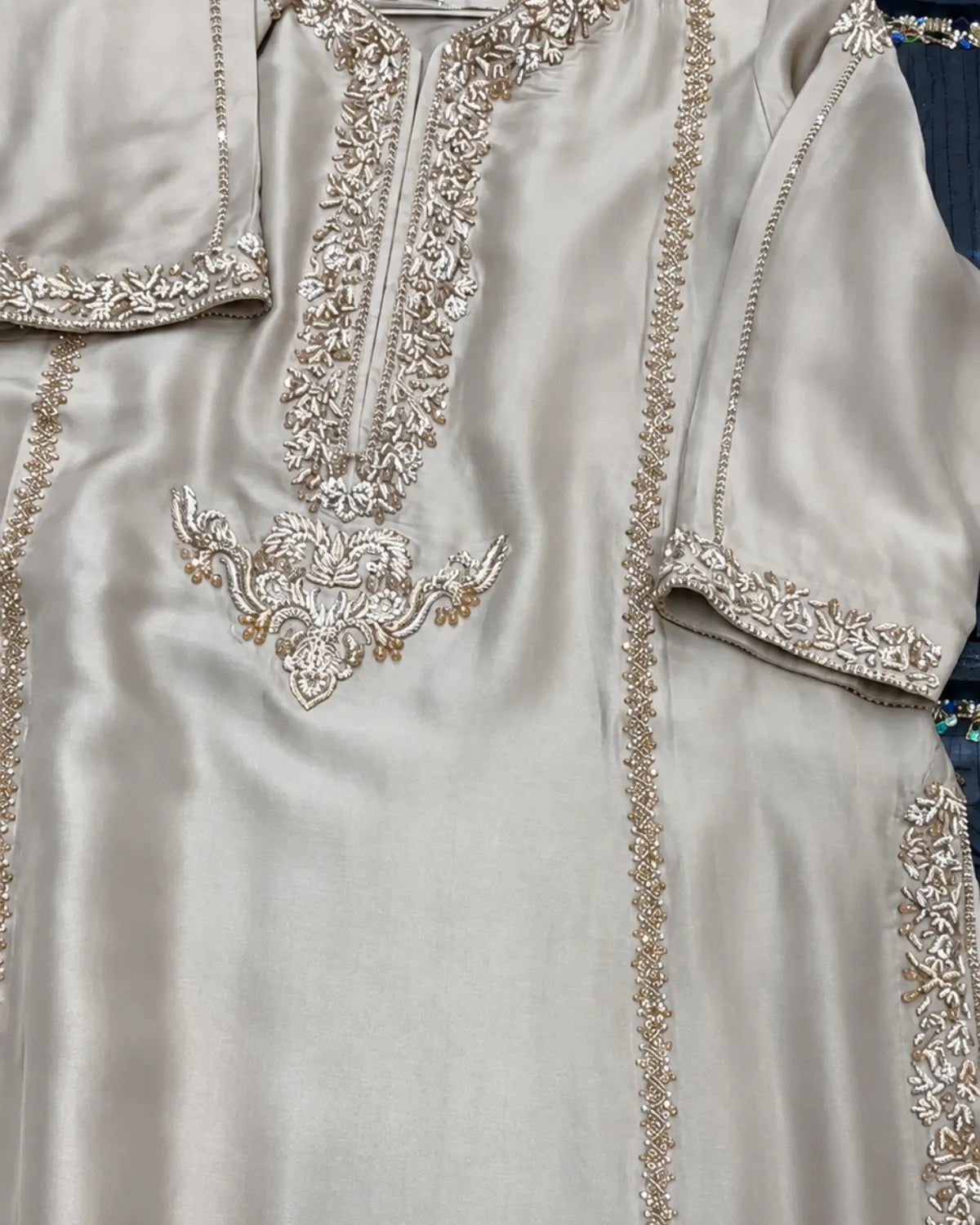 A close-up of a light beige or champagne-colored satin kurta featuring intricate silver and gold embroidery around the neckline, sleeve cuffs, and front panel.