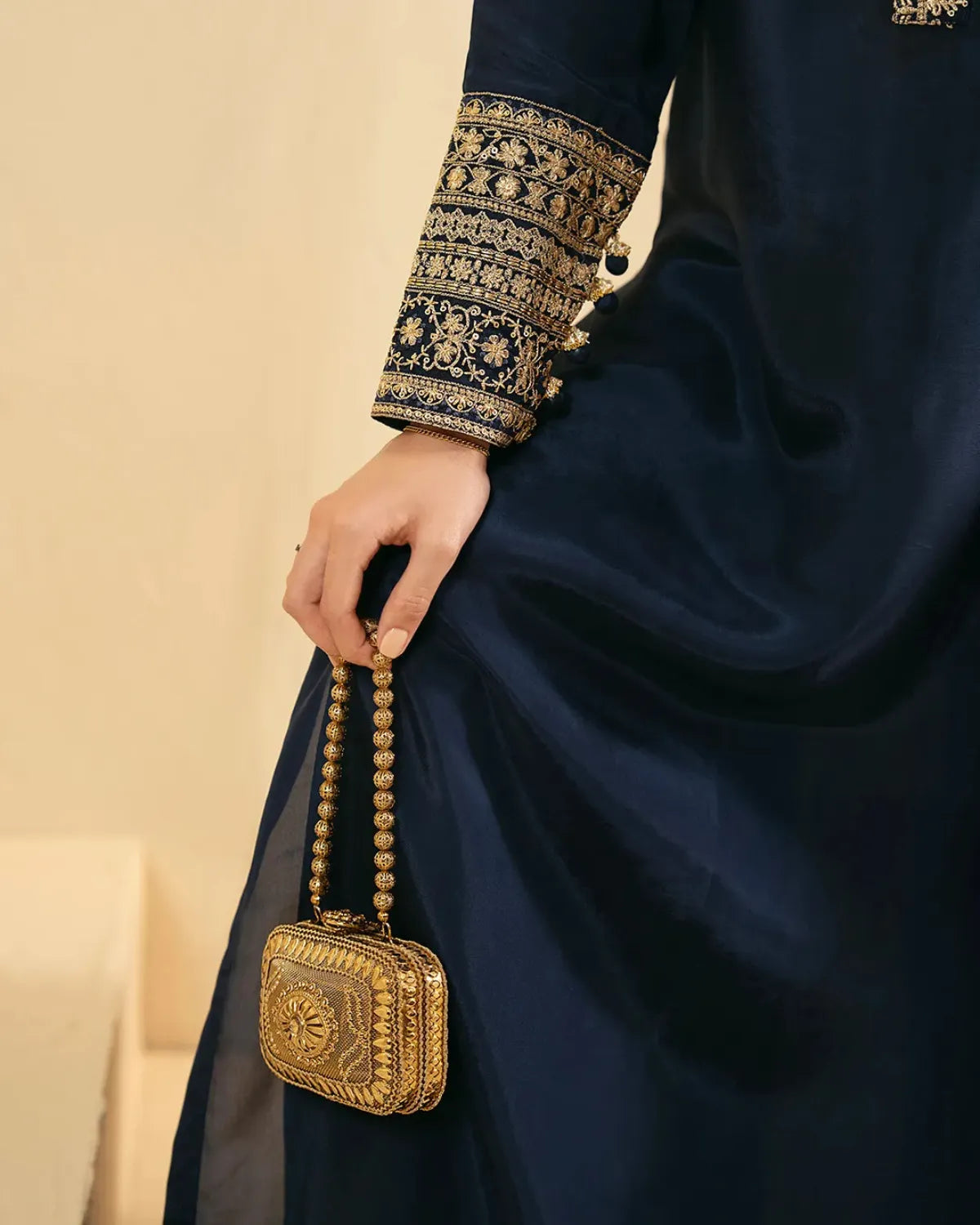 A close-up of a person wearing a dark navy satin kurta with ornate gold embroidery on the sleeve, holding a small gold metallic clutch bag with a beaded strap.