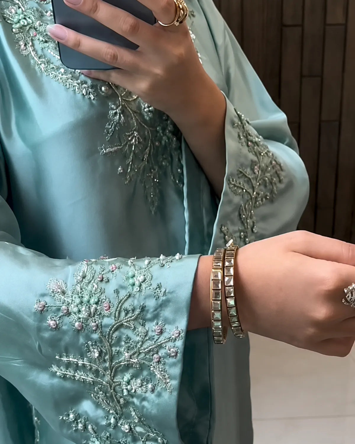 A close-up of a person wearing a light blue-green satin garment with intricate silver and pink floral embroidery on the sleeves, holding a phone in front of their face.