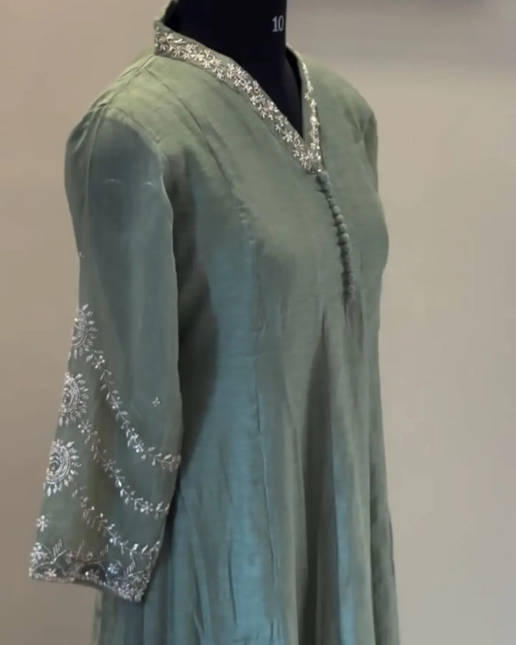 A sage green tunic or kurta displayed on a black mannequin torso, featuring silver embroidered patterns on the sleeve and a matching embroidered V-neck trim.