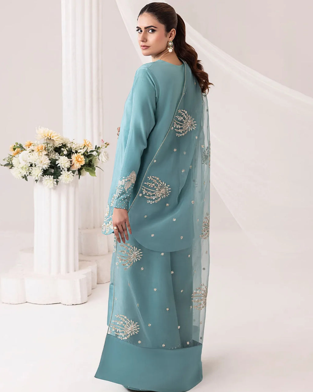 A woman modeling a teal blue-green traditional outfit with silver embroidery and a matching sheer dupatta, photographed in a bright white studio with decorative columns and flowers.
