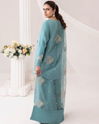 A woman modeling a teal blue-green traditional outfit with silver embroidery and a matching sheer dupatta, photographed in a bright white studio with decorative columns and flowers.