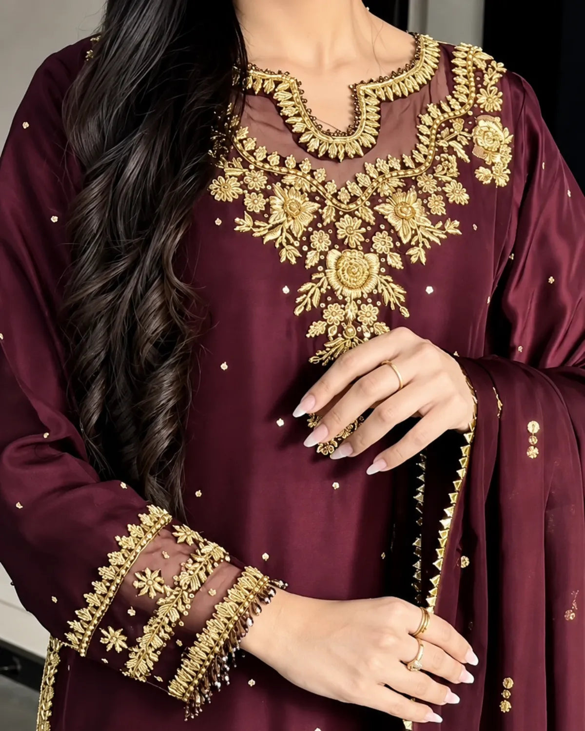 A close-up of a woman wearing a deep maroon traditional outfit with ornate gold embroidery on the neckline, sleeves, and dupatta.