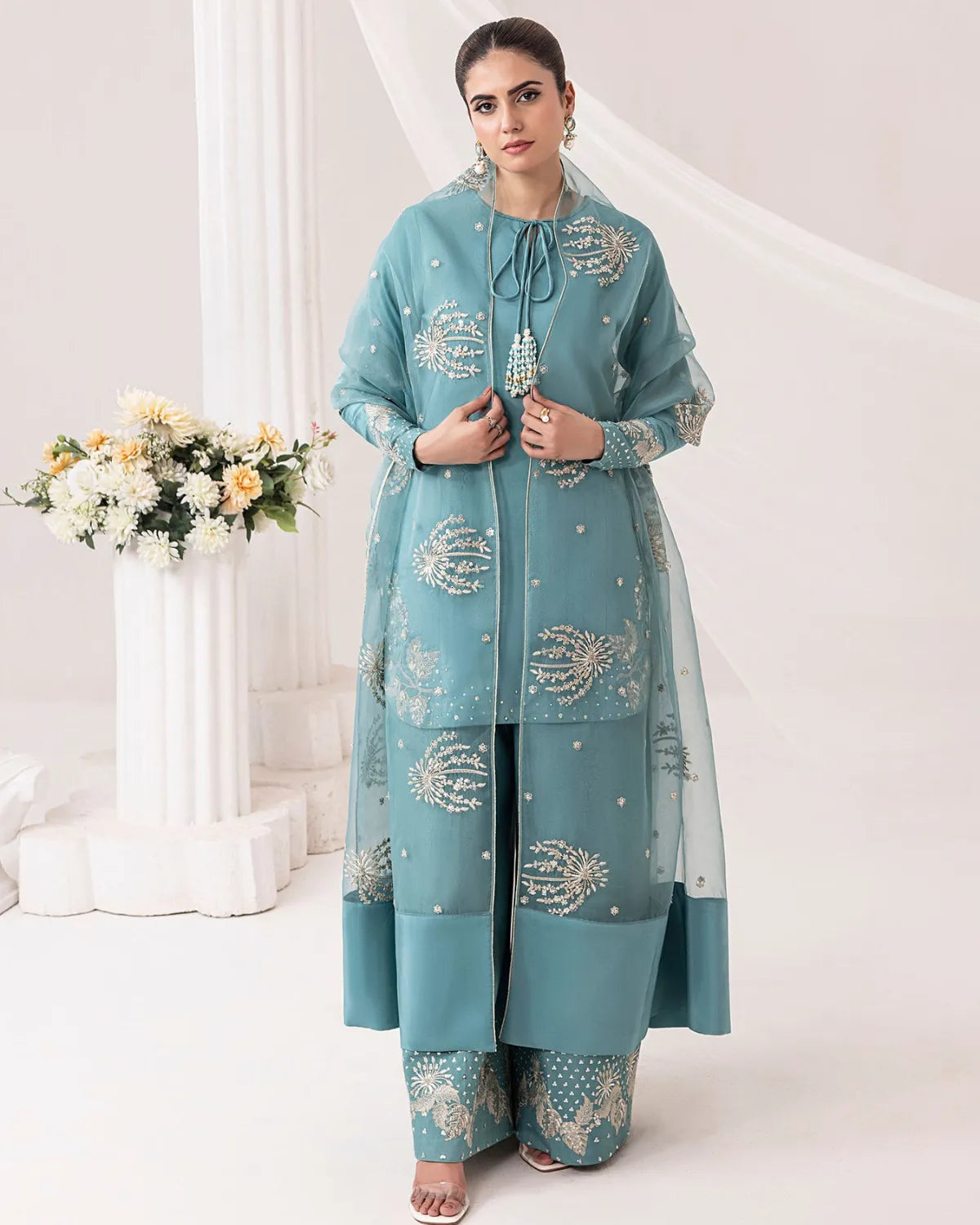 A woman modeling a traditional teal blue three-piece suit with intricate silver and gold embroidery, set against a soft, light pink and white studio backdrop with a floral arrangement on the left.