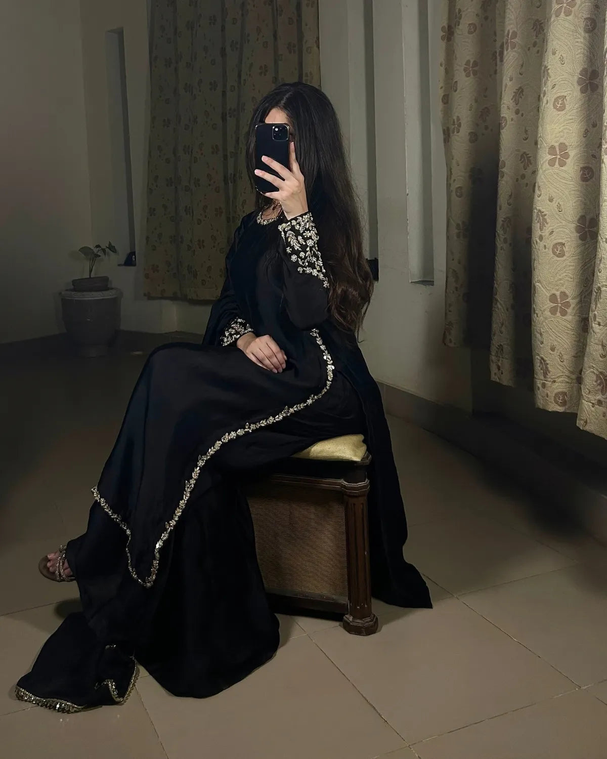 A woman wearing a black traditional outfit with silver embellished trim sits on a wooden stool in a dimly lit room with patterned curtains.