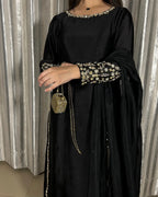 A woman wearing a black traditional outfit with gold floral embroidery on the neckline, sleeve cuffs, and along the border, holding a small gold chain bag. She is standing in front of a silver curtain backdrop.