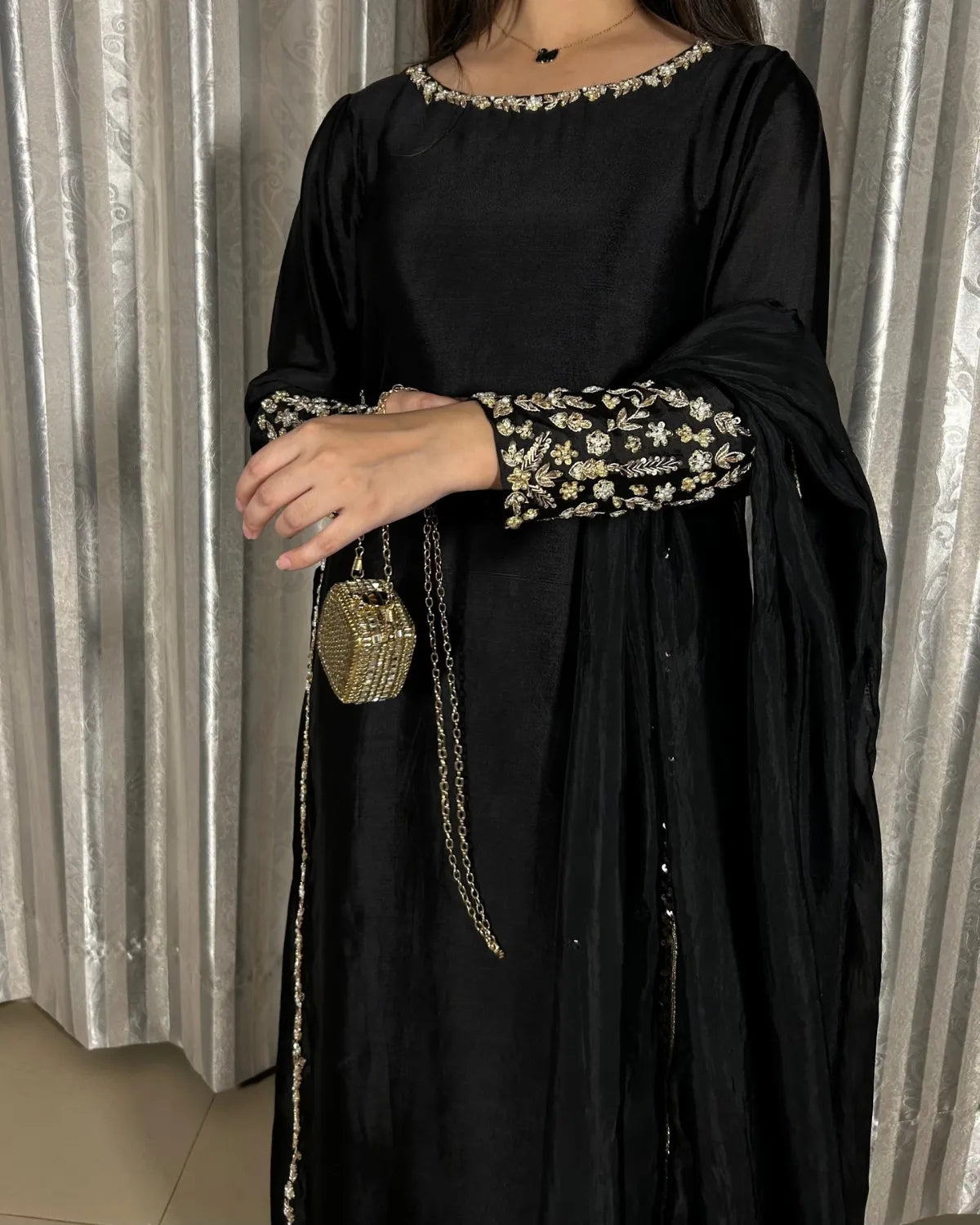 A woman wearing a black traditional outfit with gold floral embroidery on the neckline, sleeve cuffs, and along the border, holding a small gold chain bag. She is standing in front of a silver curtain backdrop.