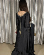 A woman viewed from behind wearing a black traditional outfit with ornate gold and silver embellishments, standing in front of silver patterned curtains.
