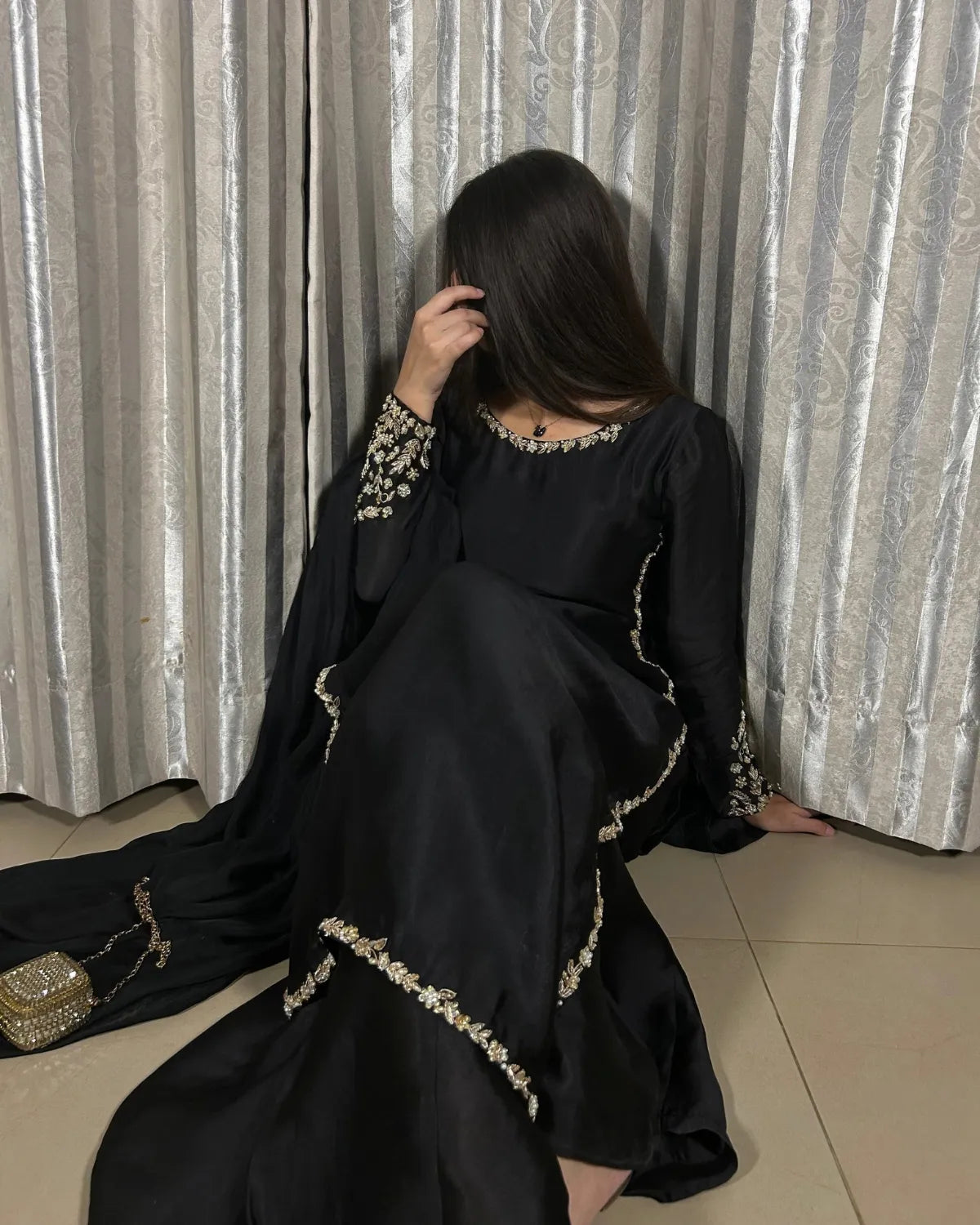 A woman wearing a black traditional dress with gold embroidered details, kneeling in front of silver patterned curtains.