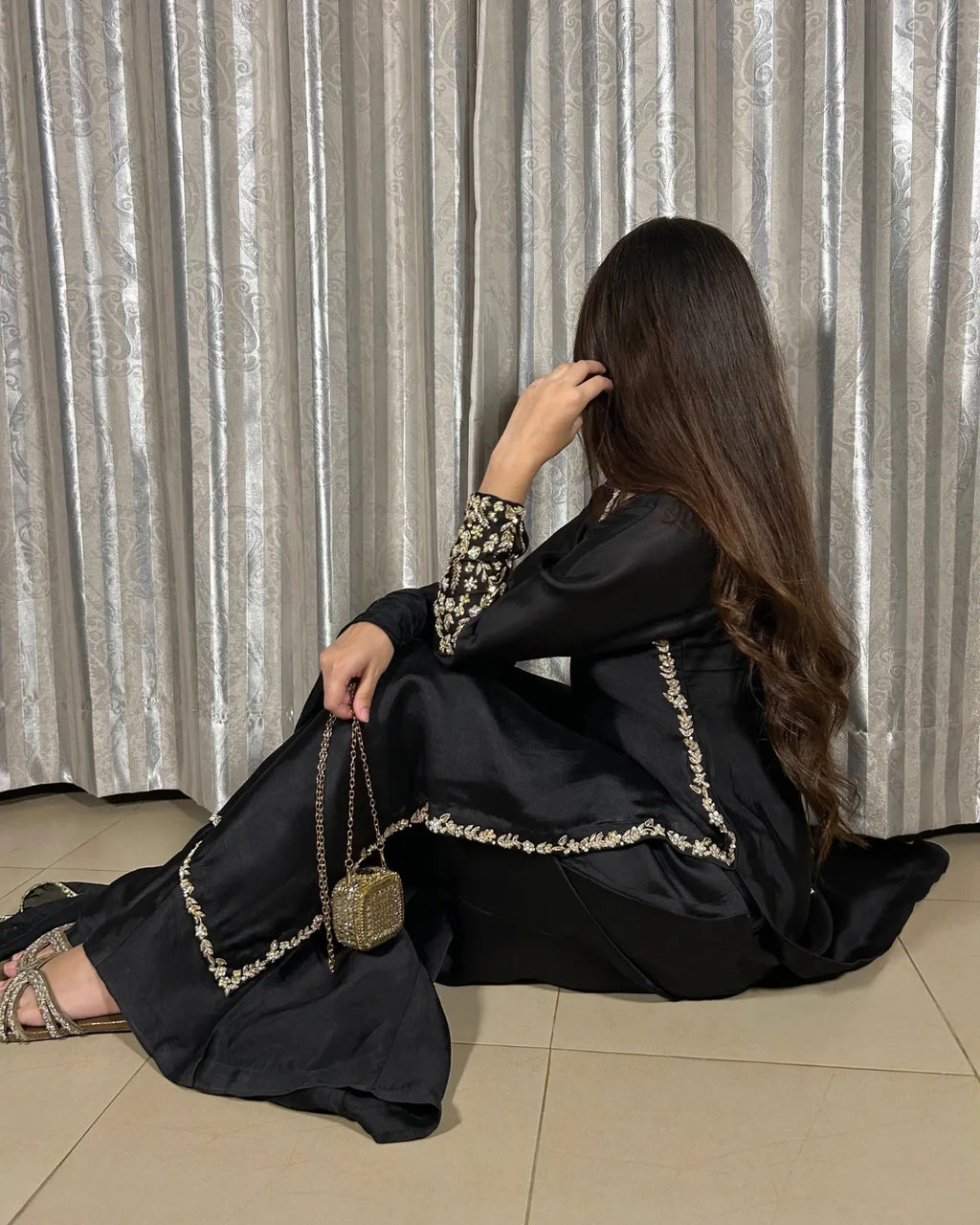A woman wearing a black abaya with gold embroidered borders sits on a tiled floor in front of silver curtains, holding a small gold chain bag.