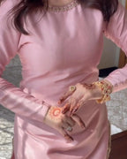 A person wearing a shiny light pink long-sleeve dress with gold embellishments at the neckline and hem, shown from the shoulders to the hips. The person has henna on their hands and is wearing gold rings and bracelets.