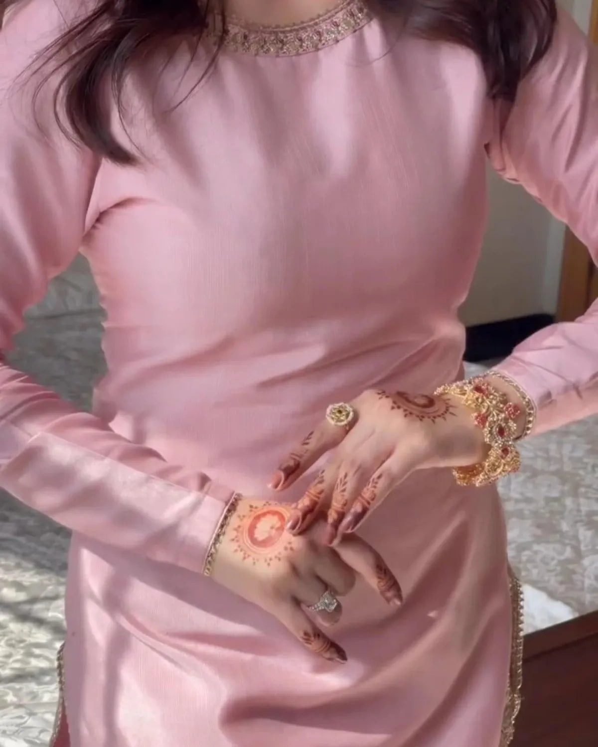 A person wearing a shiny light pink long-sleeve dress with gold embellishments at the neckline and hem, shown from the shoulders to the hips. The person has henna on their hands and is wearing gold rings and bracelets.