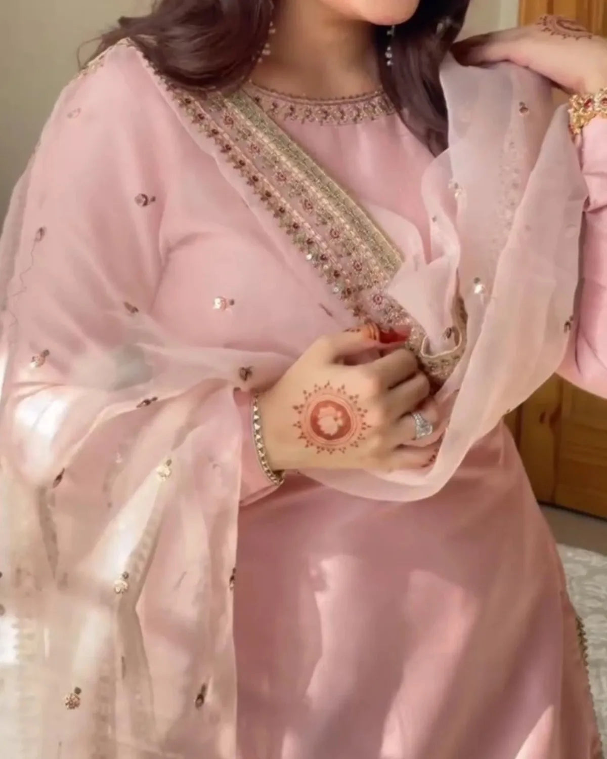 A woman wearing a light pink traditional outfit with a sheer matching dupatta, decorated with gold and silver mirror work and sequins along the neckline. She is shown from the chest up in an indoor setting.