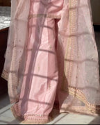 A light pink traditional outfit with matching sheer dupatta draped over the body, featuring gold embroidered borders and small gold motifs, shown in warm natural sunlight with window-frame shadows.