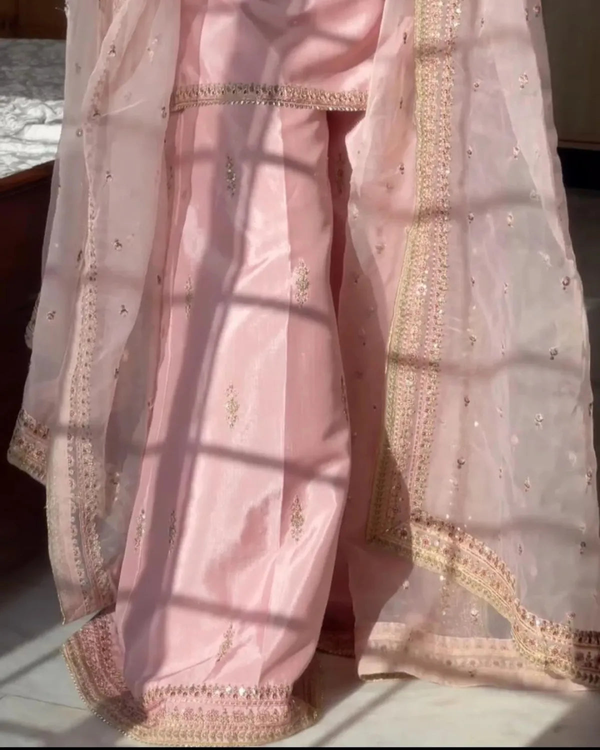 A light pink traditional outfit with matching sheer dupatta draped over the body, featuring gold embroidered borders and small gold motifs, shown in warm natural sunlight with window-frame shadows.
