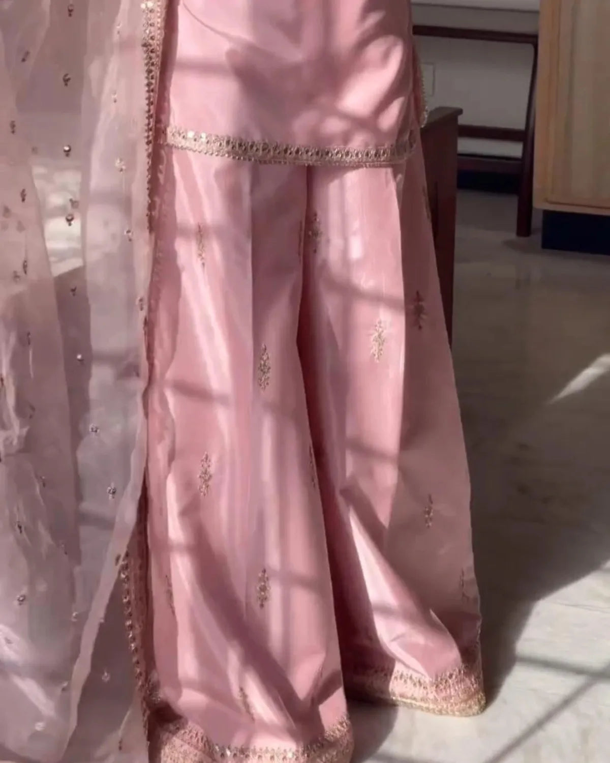 A close-up of a pink traditional outfit with wide flared pants, embroidered details, and a matching sheer dupatta draped over one leg, photographed indoors with sunlight and shadows.