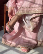 A person wearing a light pink traditional outfit with heavily embroidered borders and matching embellished flat shoes, seated indoors with sunlight casting shadows across the fabric.