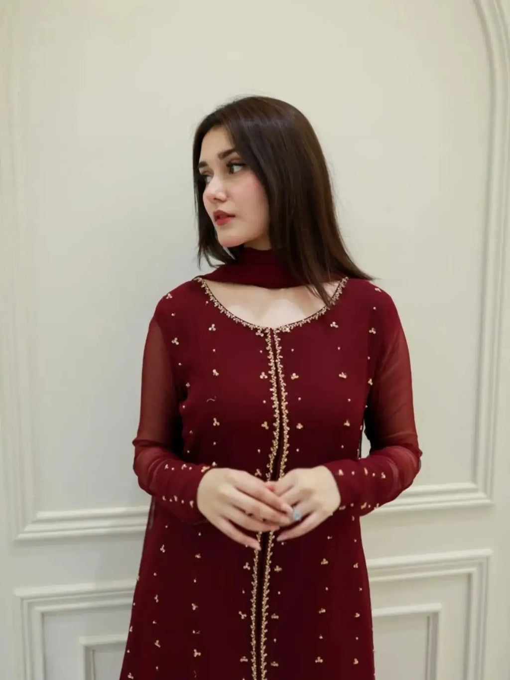 A woman wearing a deep maroon long-sleeve kurta with gold embroidery and a matching neck scarf, standing against a white paneled wall.